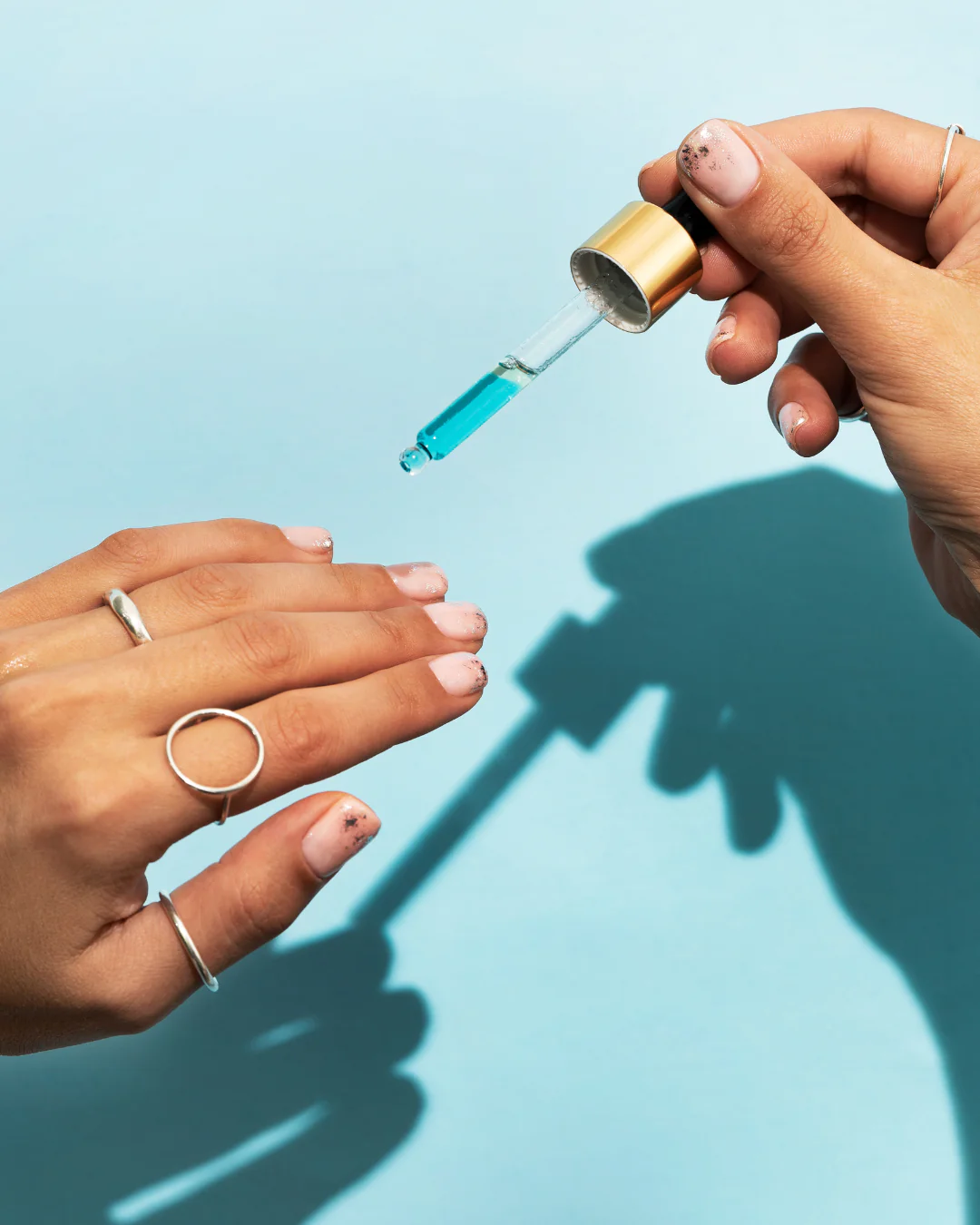 hands with glitter nails using a glass dropper to apply blue cuticle oil
