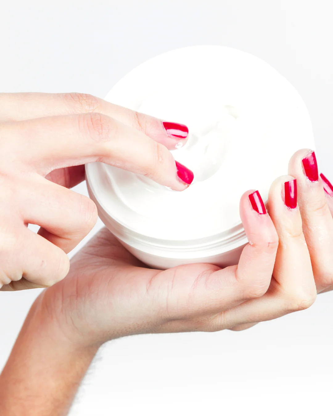 person with bright red nails scooping white moisturizing cream from a large tub