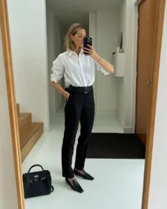 woman in a white button-down shirt and black trousers taking a mirror selfie.