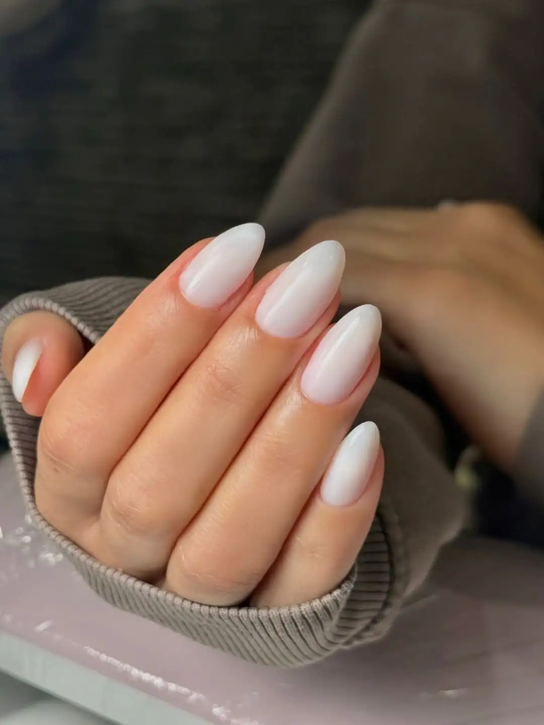 glossy milky white almond nails peeking out from a ribbed gray sweater sleeve.
