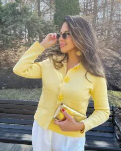 woman wearing a butter yellow cardigan with floral buttons and white trousers outdoors.