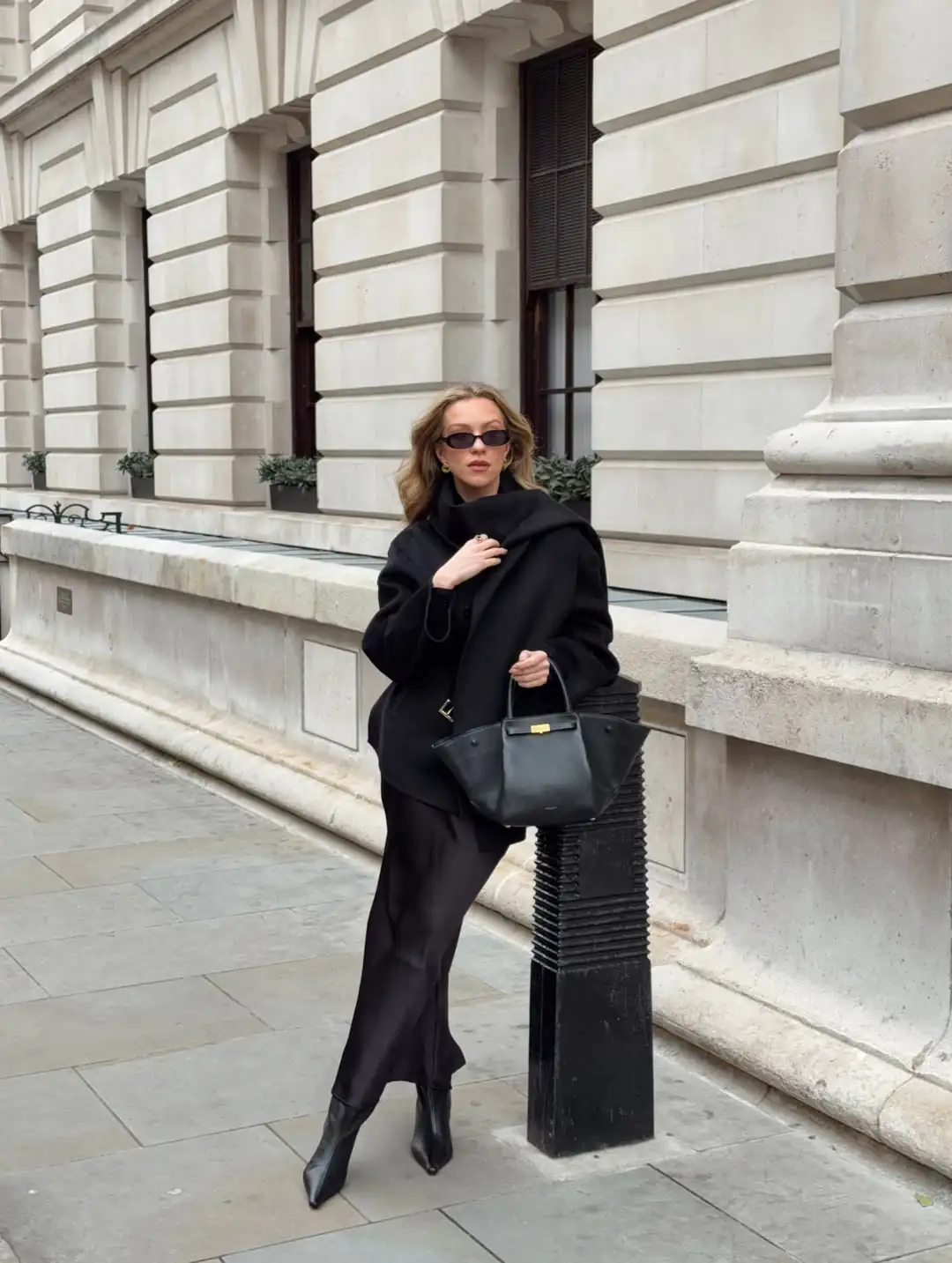 woman in black wrap coat and silk midi dress leaning on street post