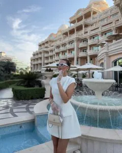 woman in a white mini dress and sheer gloves standing by a fountain in dubai.