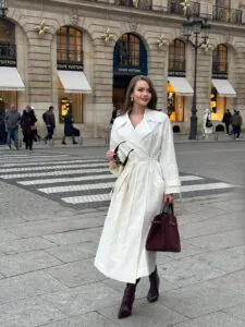 woman in a long white trench coat and dark boots walking near luxury storefronts.