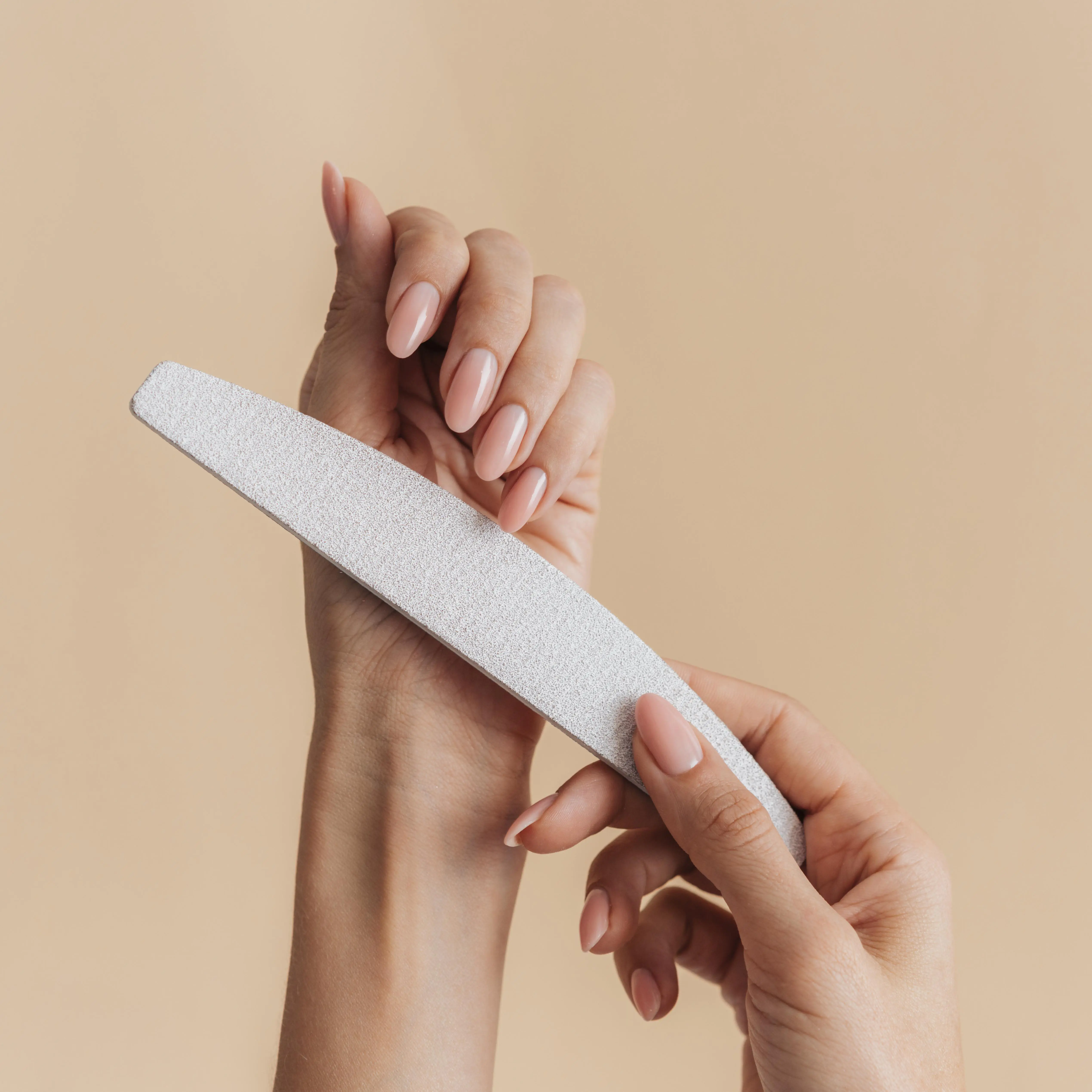 woman with a glossy nude manicure holding a gray nail buffer against a tan background