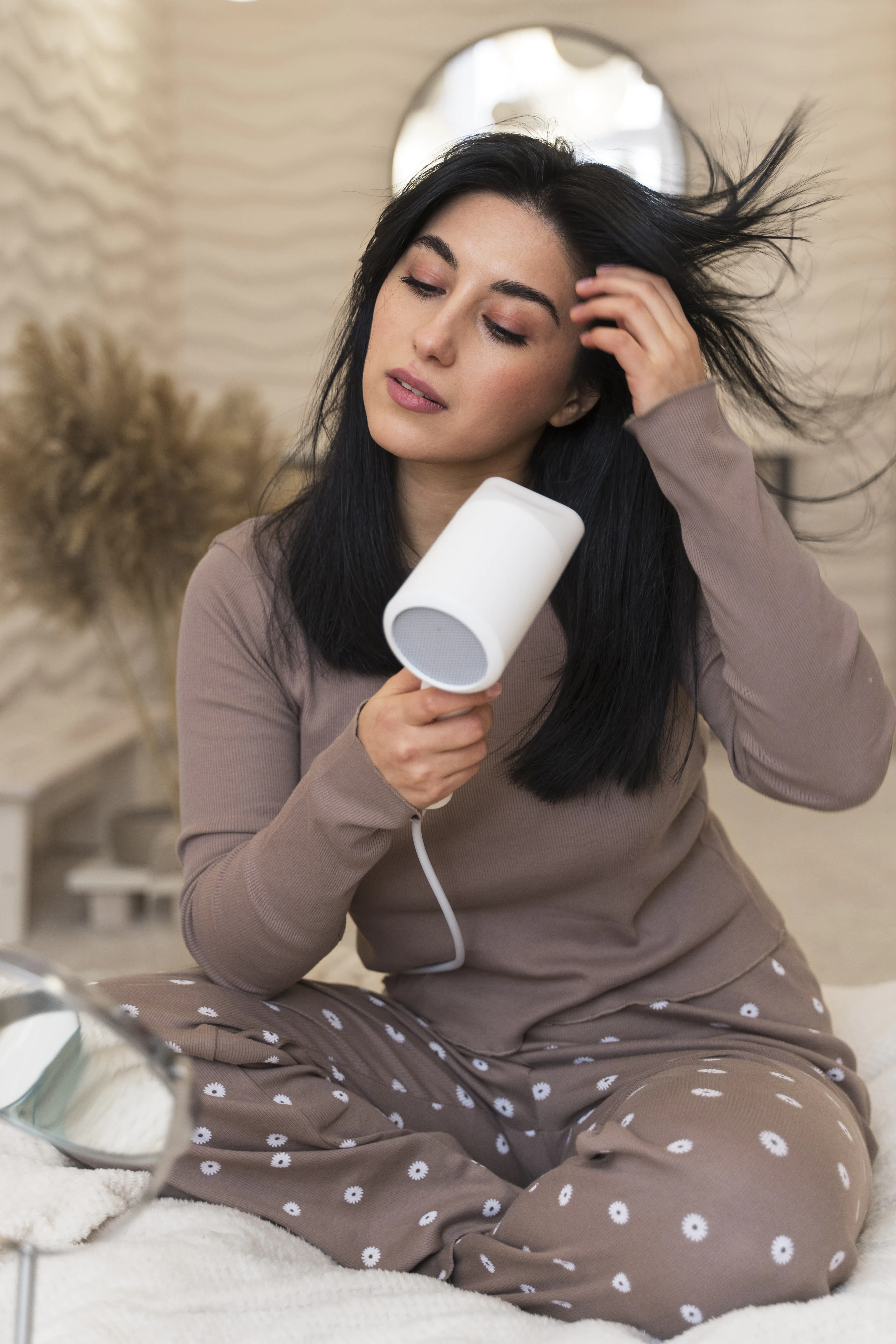 woman sitting on a bed using a white hair dryer on her dark hair