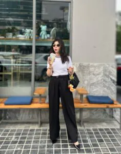 woman in white t-shirt and black wide-leg trousers drinking iced coffee at a cafe.