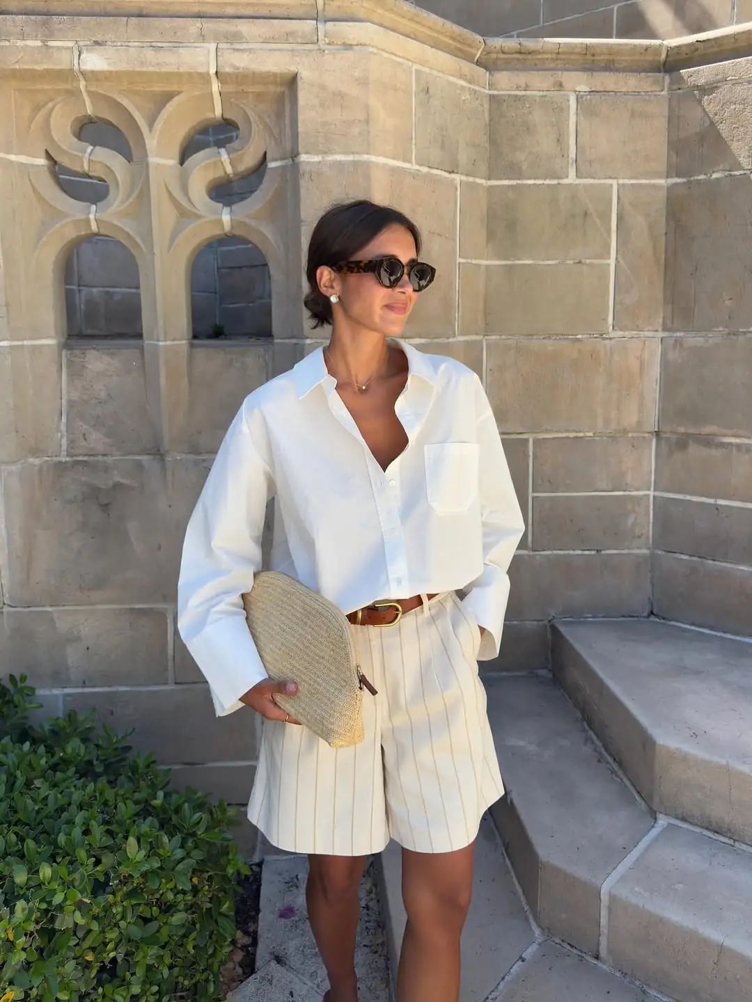 woman wearing oversized white shirt and striped linen shorts holding a woven clutch