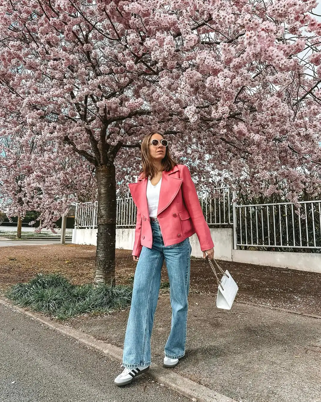 woman in pink coat and jeans standing under blooming cherry blossom tree
