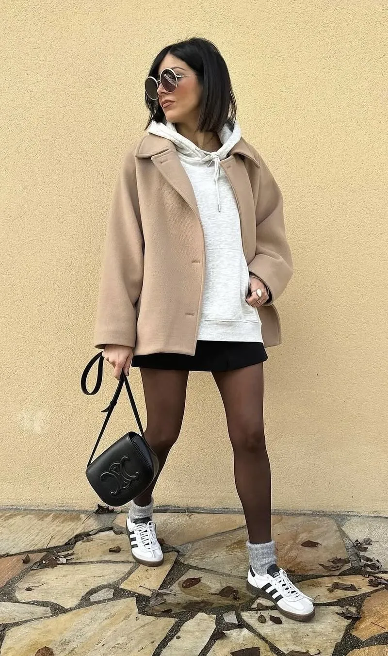 woman in camel coat and grey hoodie standing against yellow wall