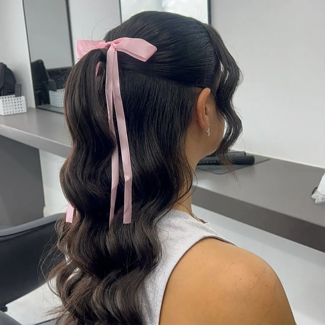 brunette woman with a wavy half-up half-down hairstyle tied with a pink ribbon bow.