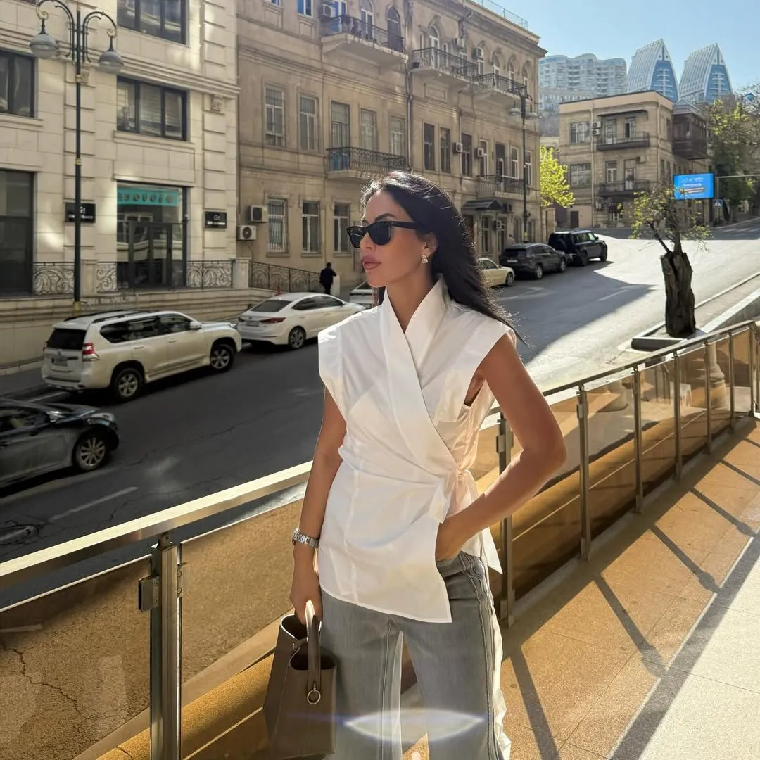 woman wearing sleeveless white wrap top and sunglasses standing on a sunny city balcony