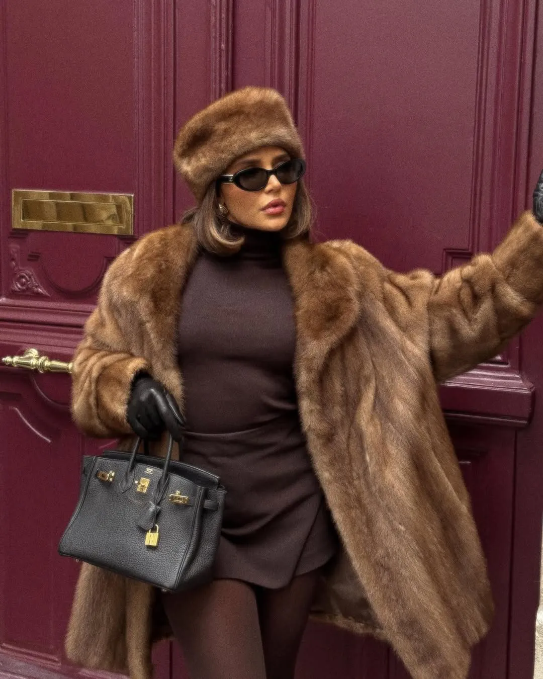 woman in brown fur coat and matching hat posing against a dark red door