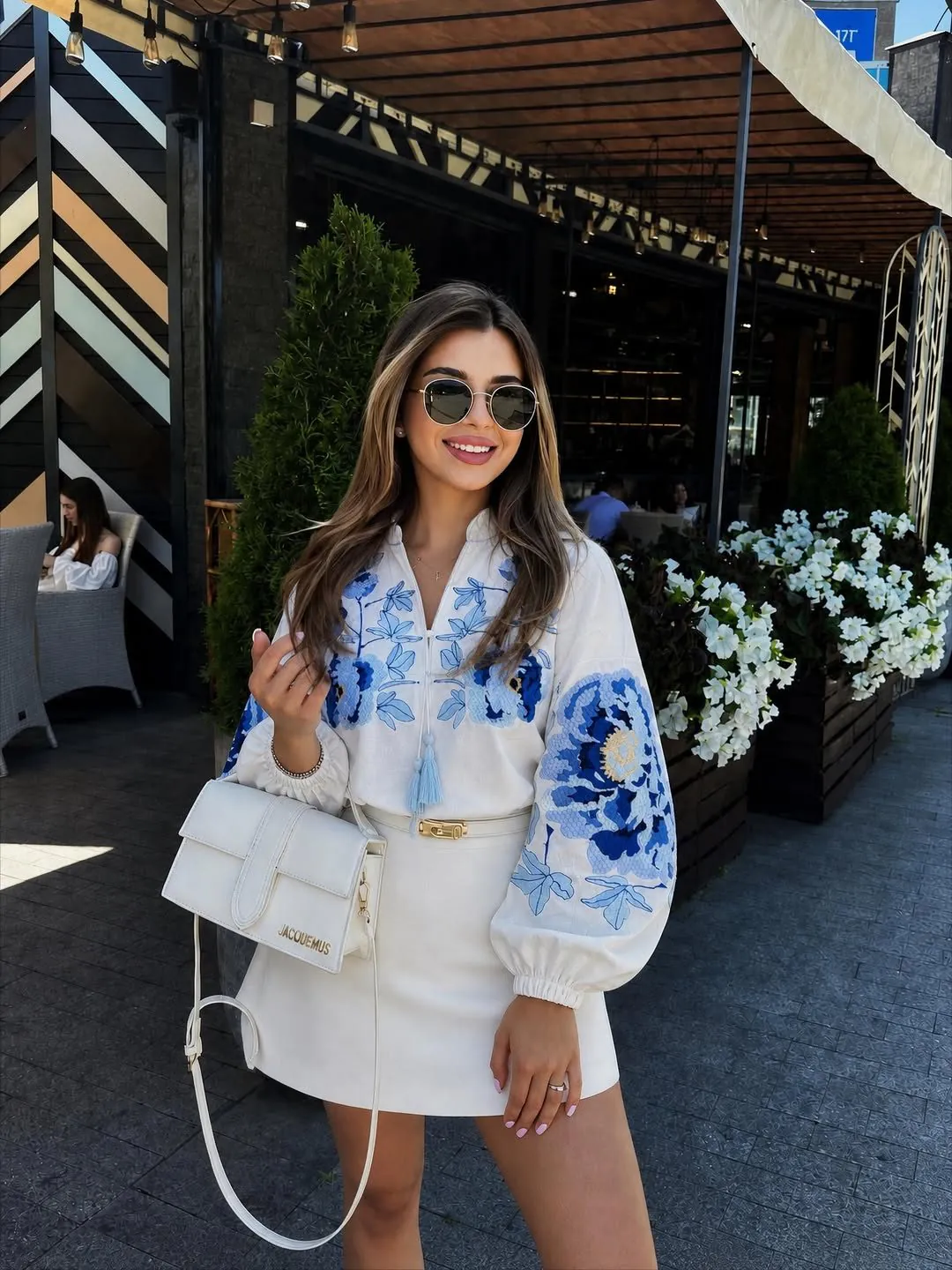 woman wearing white embroidered blouse and white skirt holding a luxury handbag outdoors