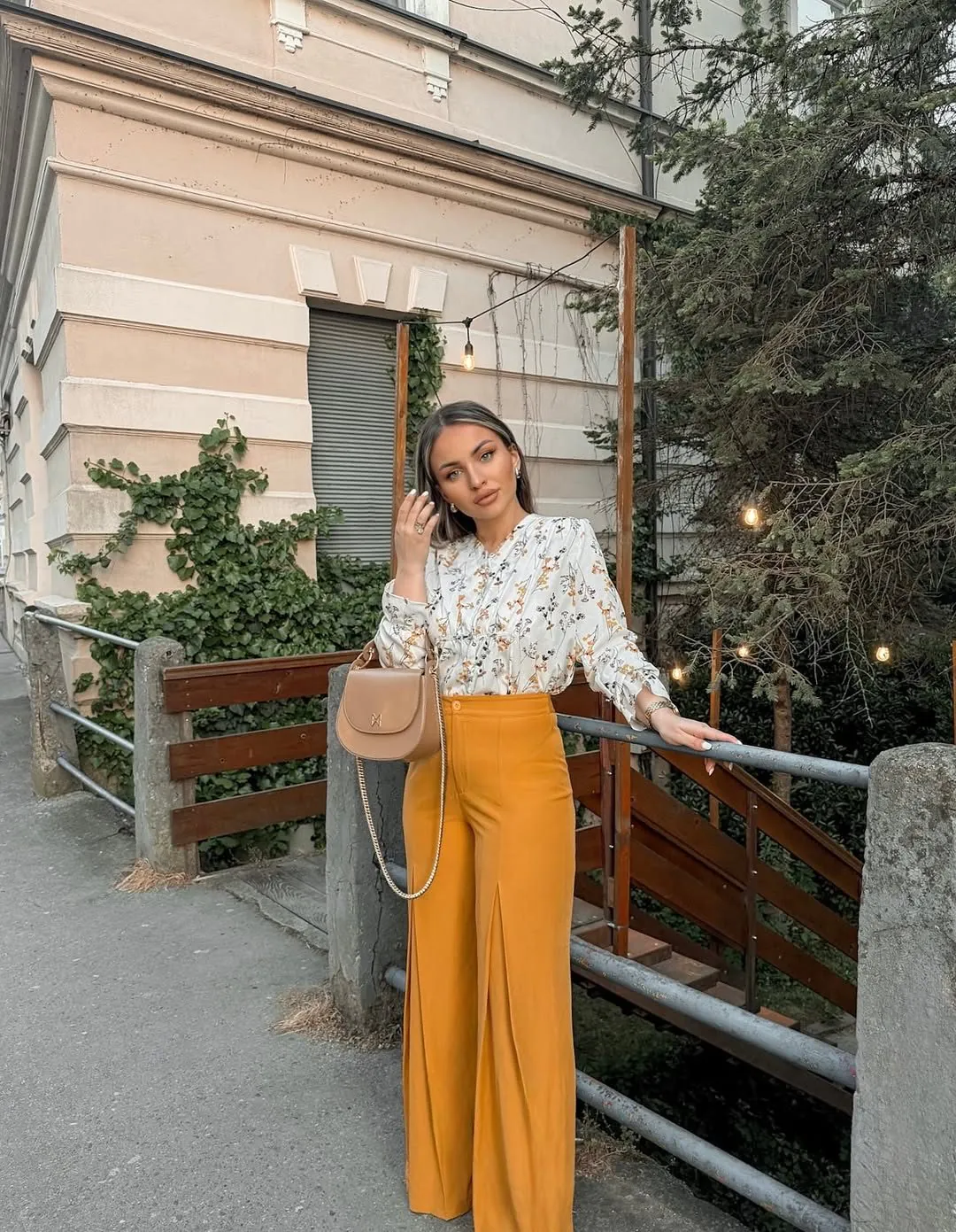 woman in floral blouse and mustard yellow wide-leg trousers standing by a wooden fence