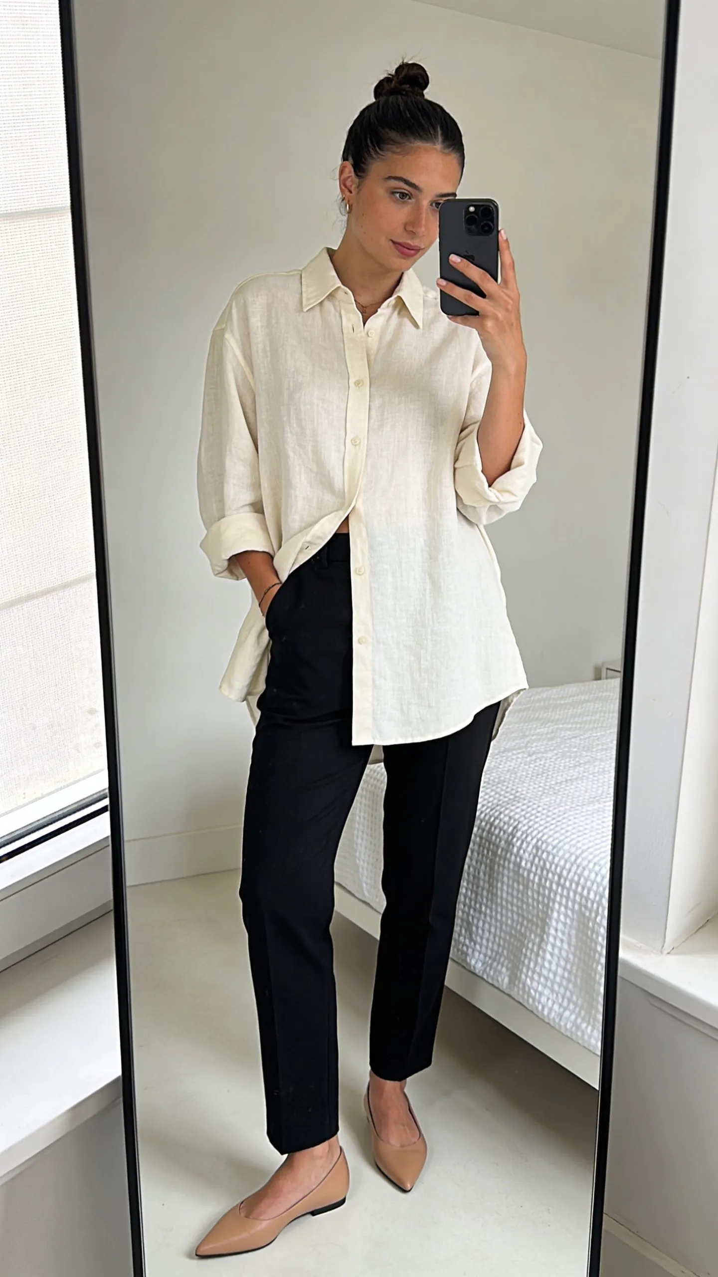 woman in oversized cream linen shirt and black trousers taking a bedroom mirror selfie