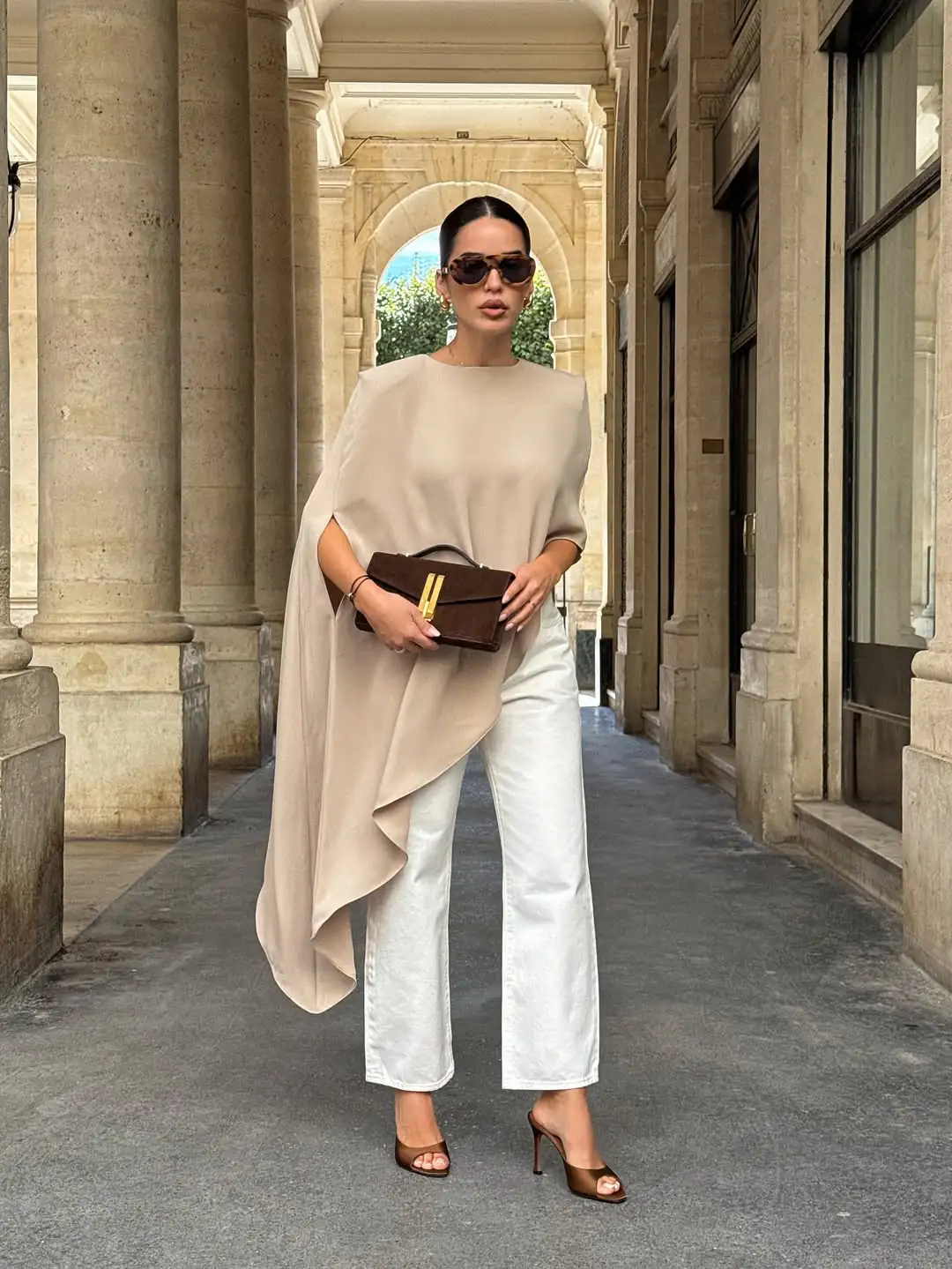 woman wearing tan asymmetrical cape top and white jeans standing in stone archway