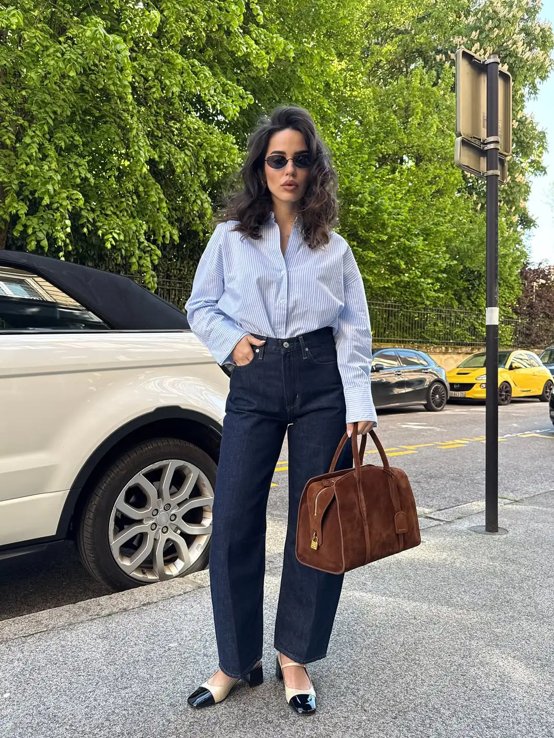 woman in striped button down shirt and dark wash jeans holding brown suede bag