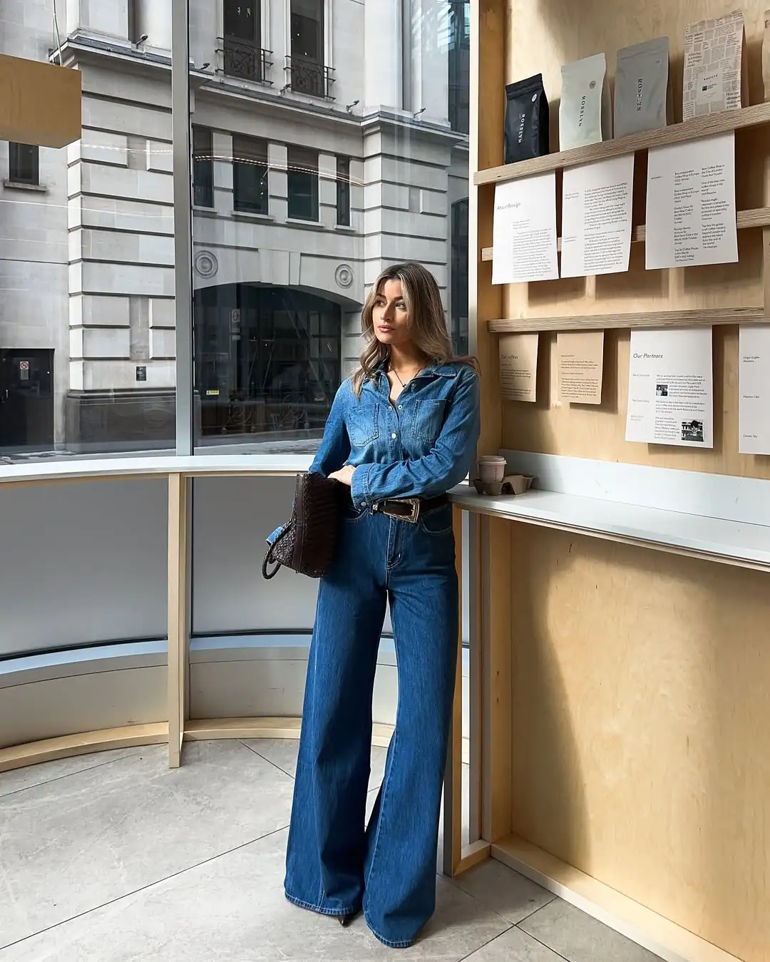 woman wearing denim shirt and wide leg jeans leaning against wooden cafe wall