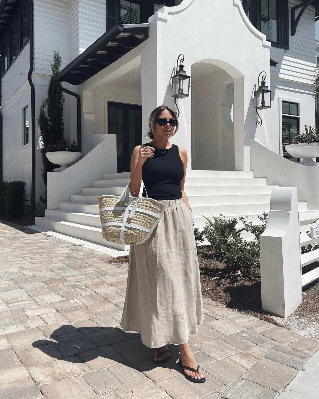 woman in black tank top and beige linen maxi skirt holding a straw bag.