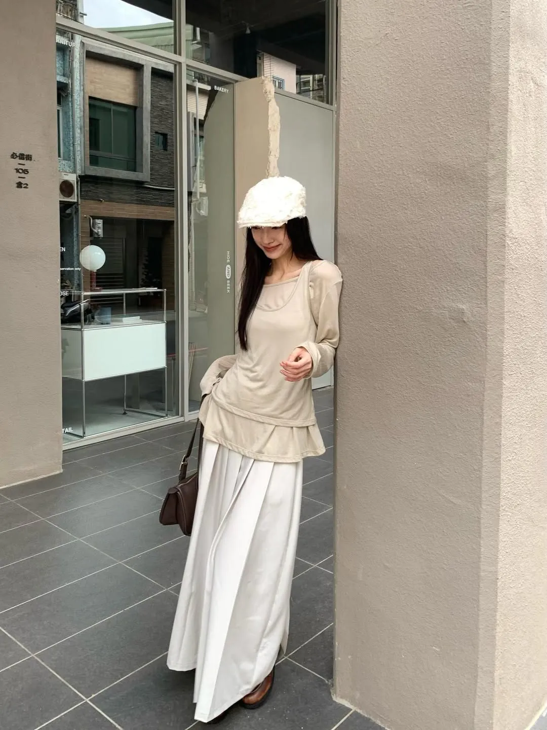 woman in beige long sleeve top and white wide-leg skirt leaning against a wall.