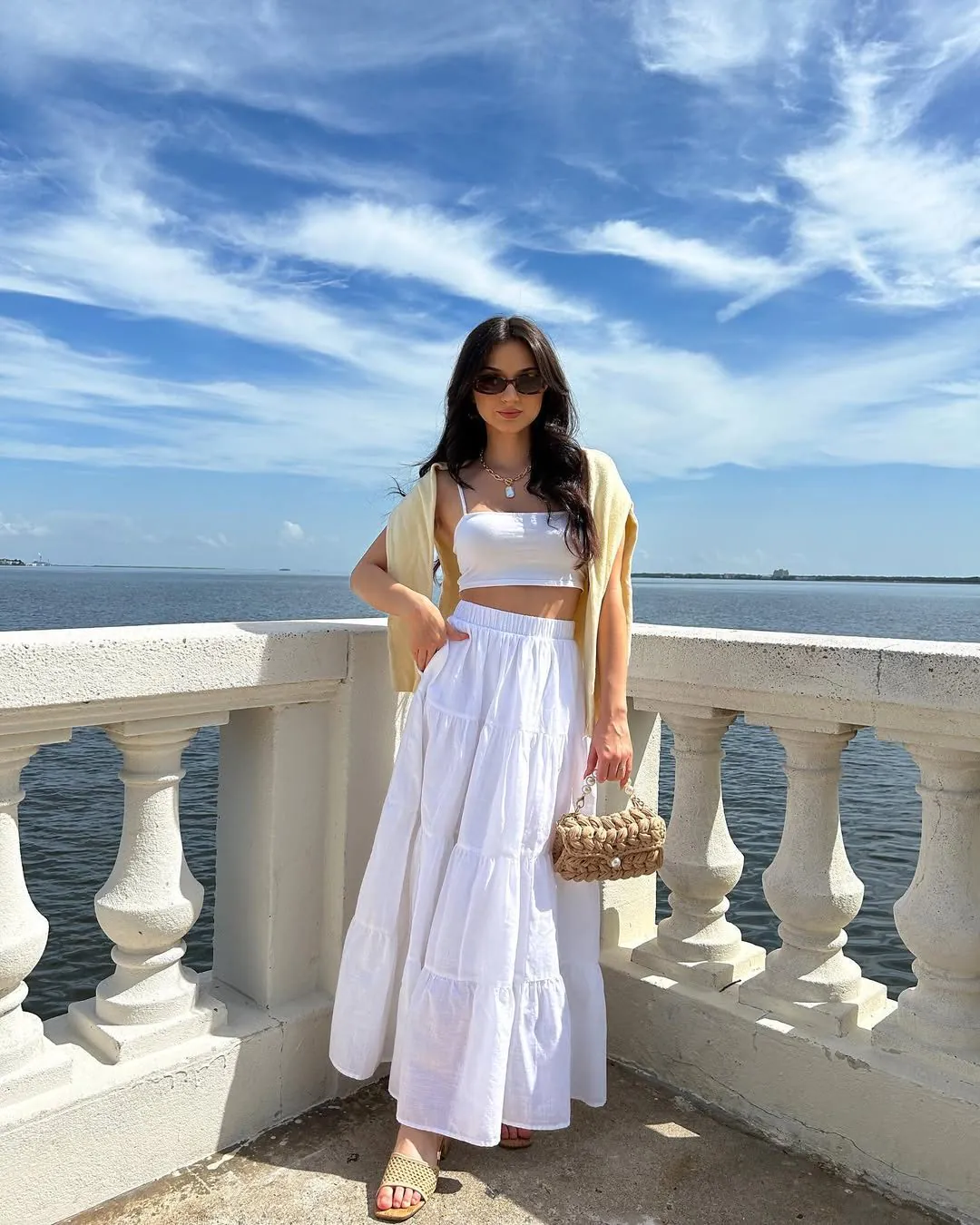 woman wearing white crop top and tiered maxi skirt standing by a coastal balcony.