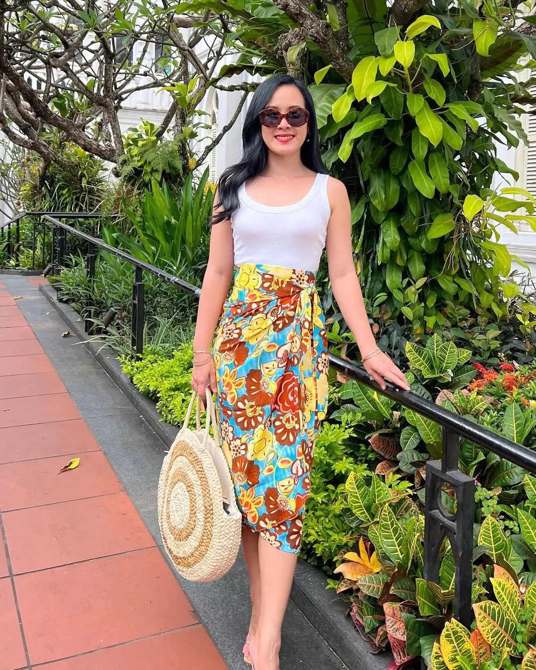 woman in white tank top and vibrant tropical floral sarong skirt with straw tote.