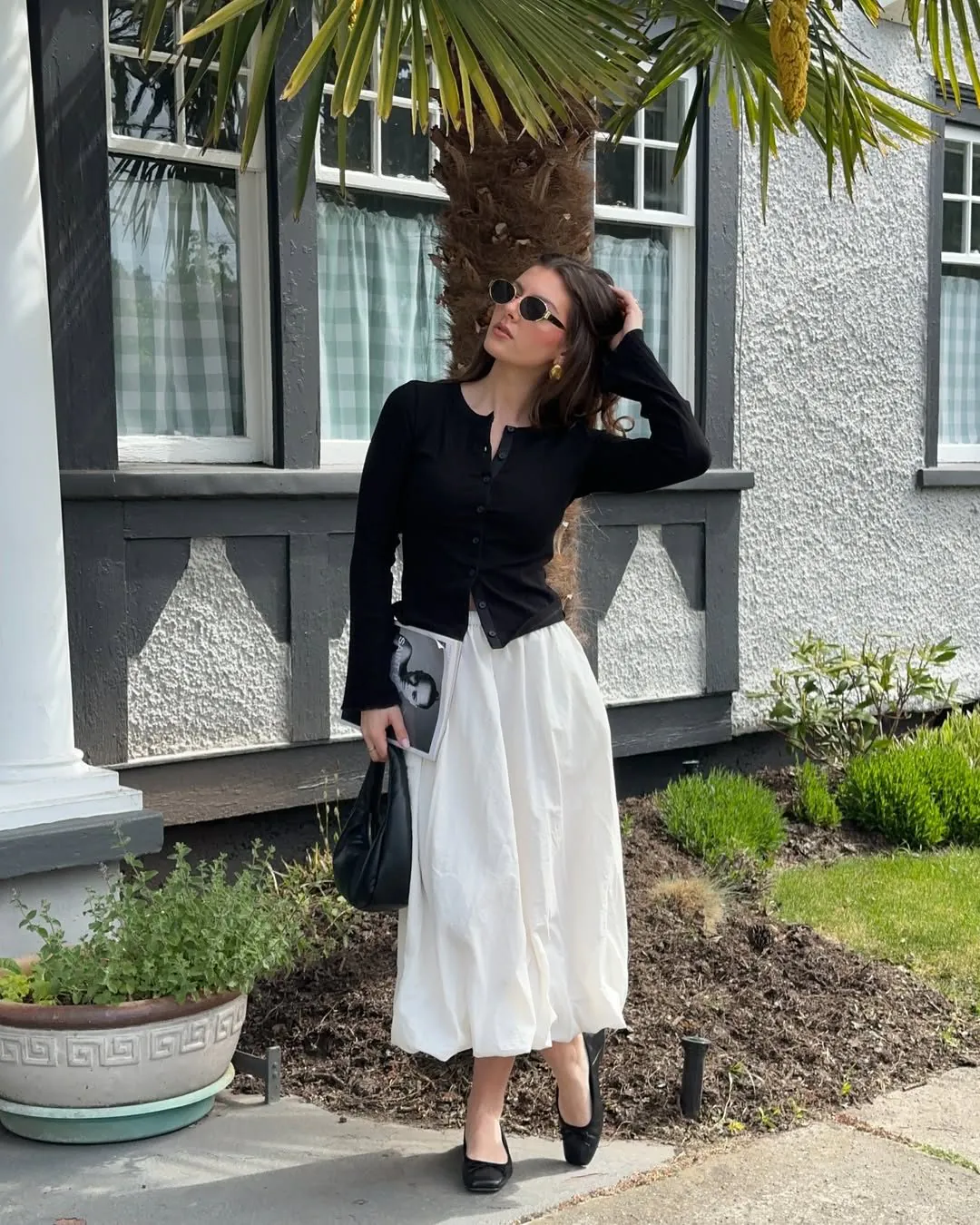 woman in black cardigan and long white flowy skirt holding a black handbag.