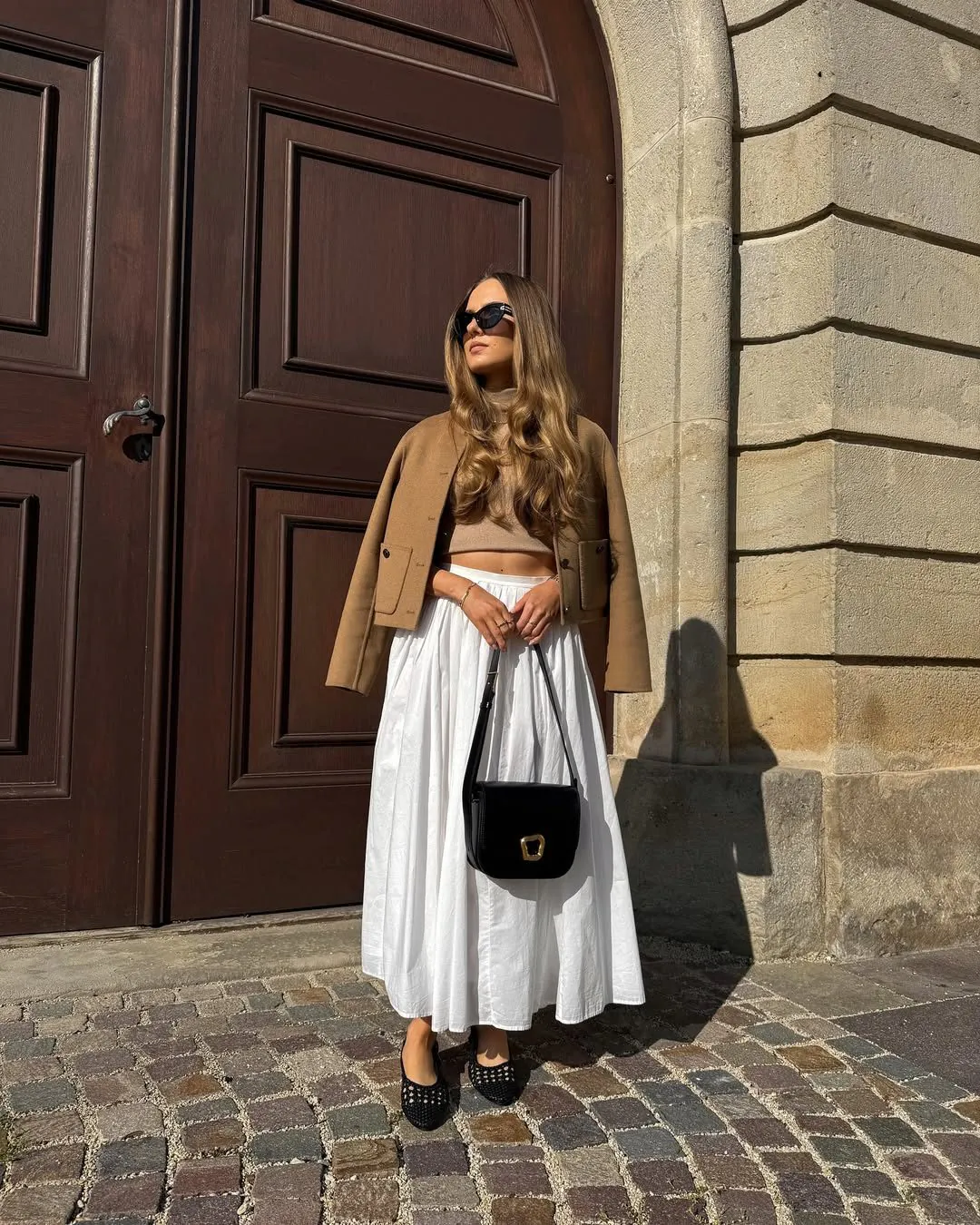 woman wearing tan cropped jacket and long white pleated skirt with black handbag.