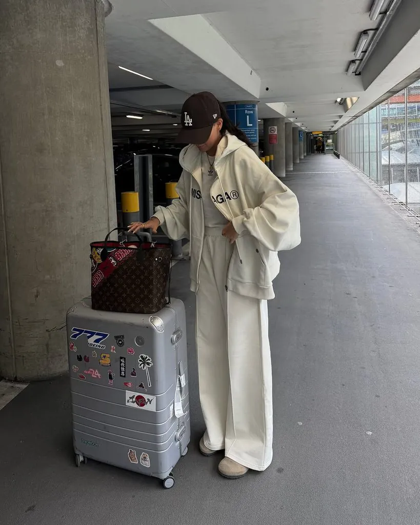 woman in cream oversized tracksuit and brown baseball cap beside silver luggage