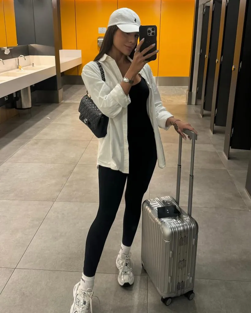 woman wearing black leggings white shirt and cap standing next to silver suitcase