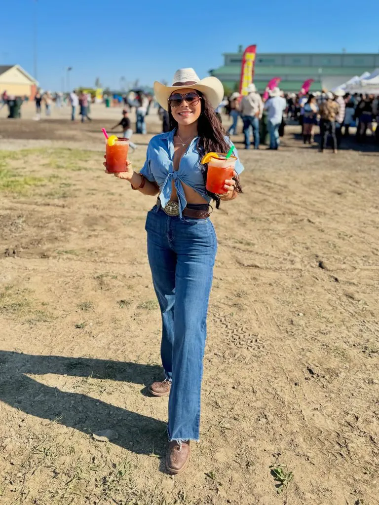 woman wearing a white cowboy hat and tied denim shirt holding two red drinks