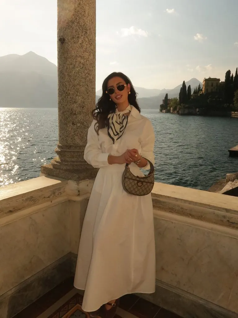 woman in a white dress and neck scarf standing by a balcony overlooking the water