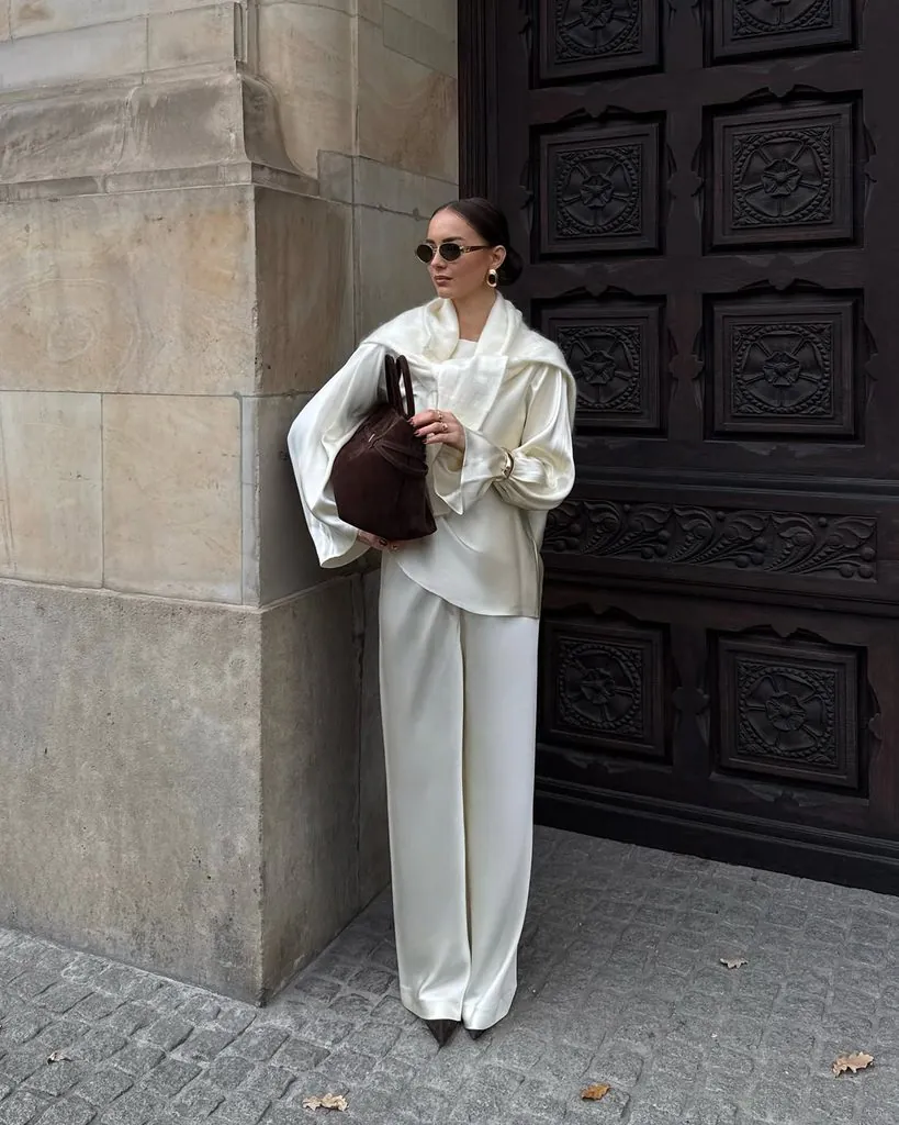 woman in an elegant all-white outfit holding a brown handbag near a carved wooden door