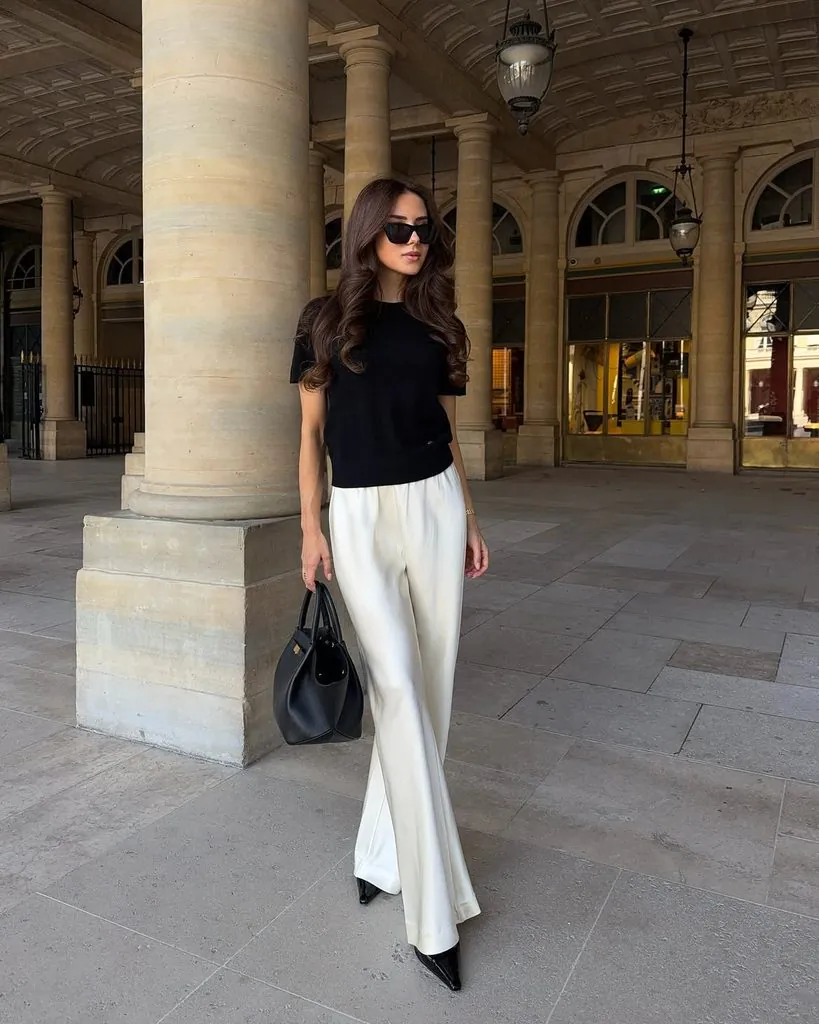 woman wearing a black short-sleeved top and white wide-leg trousers standing near stone pillars