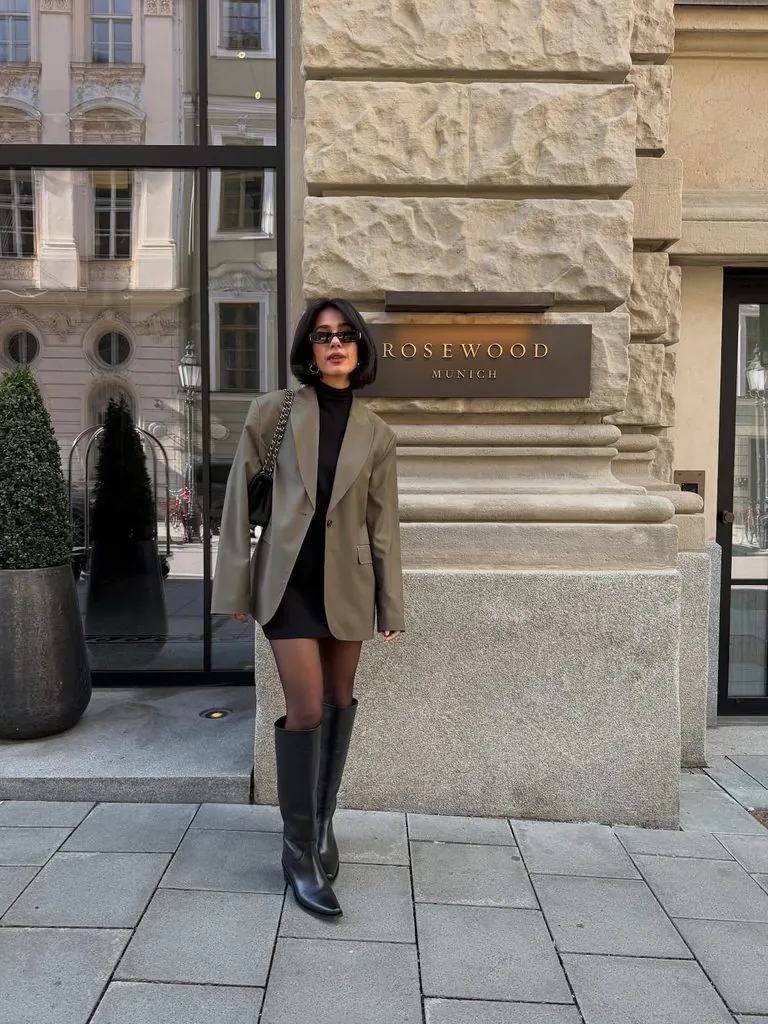 woman wearing an olive blazer black mini dress and knee-high leather boots by hotel sign