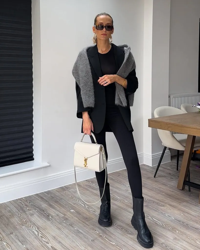 woman in black leggings, blazer, grey shoulder sweater, holding white handbag indoors