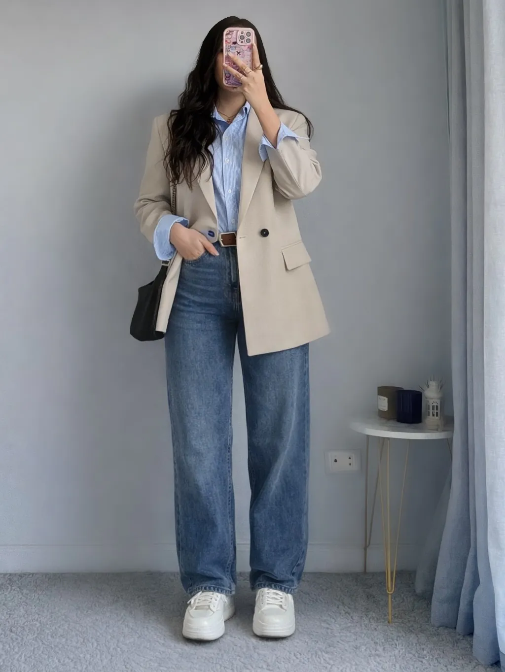 woman in a beige blazer, blue striped shirt, and wide-leg jeans taking a selfie