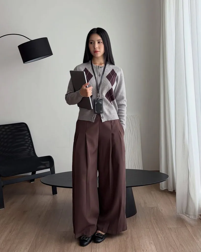 woman wearing a grey argyle cardigan and brown wide-leg trousers while holding a laptop