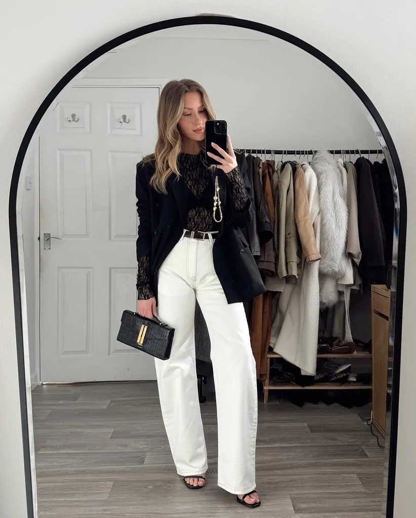 woman taking mirror selfie wearing black blazer, lace top, and white jeans