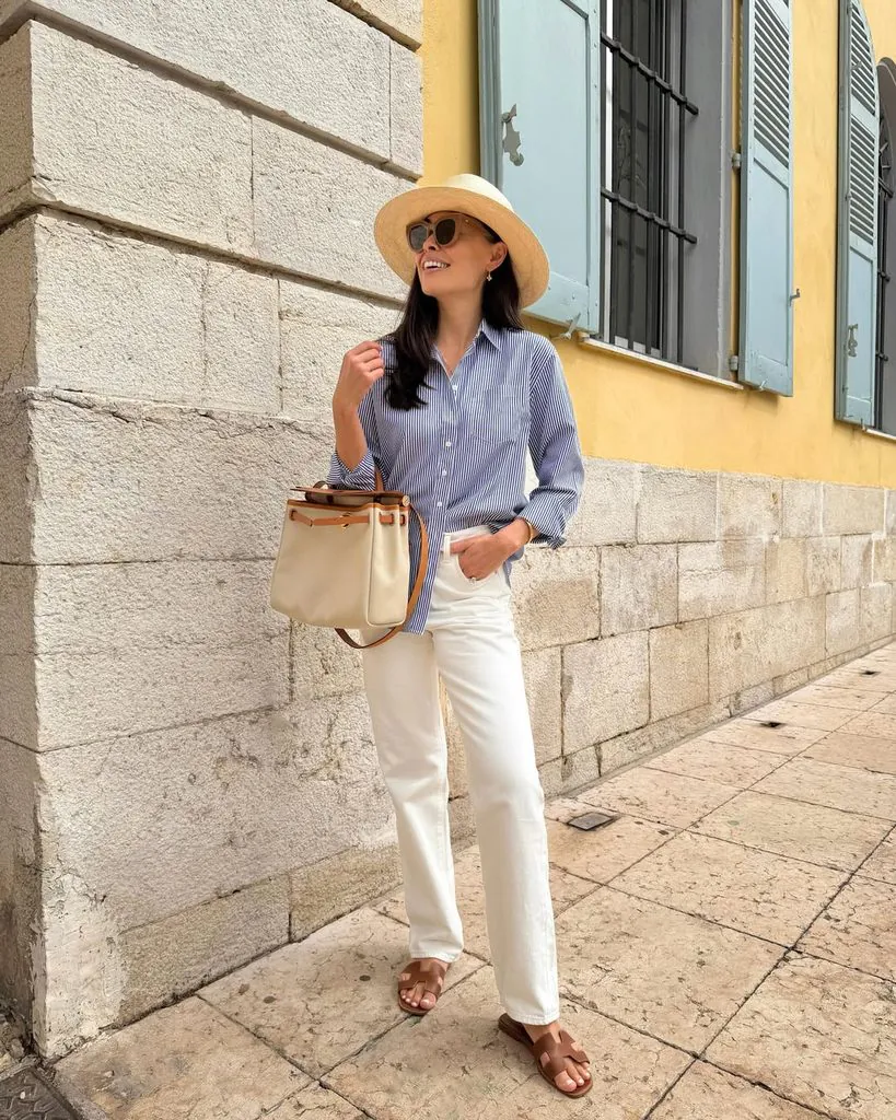 smiling woman wearing a blue striped button-down shirt, straw hat, and white jeans