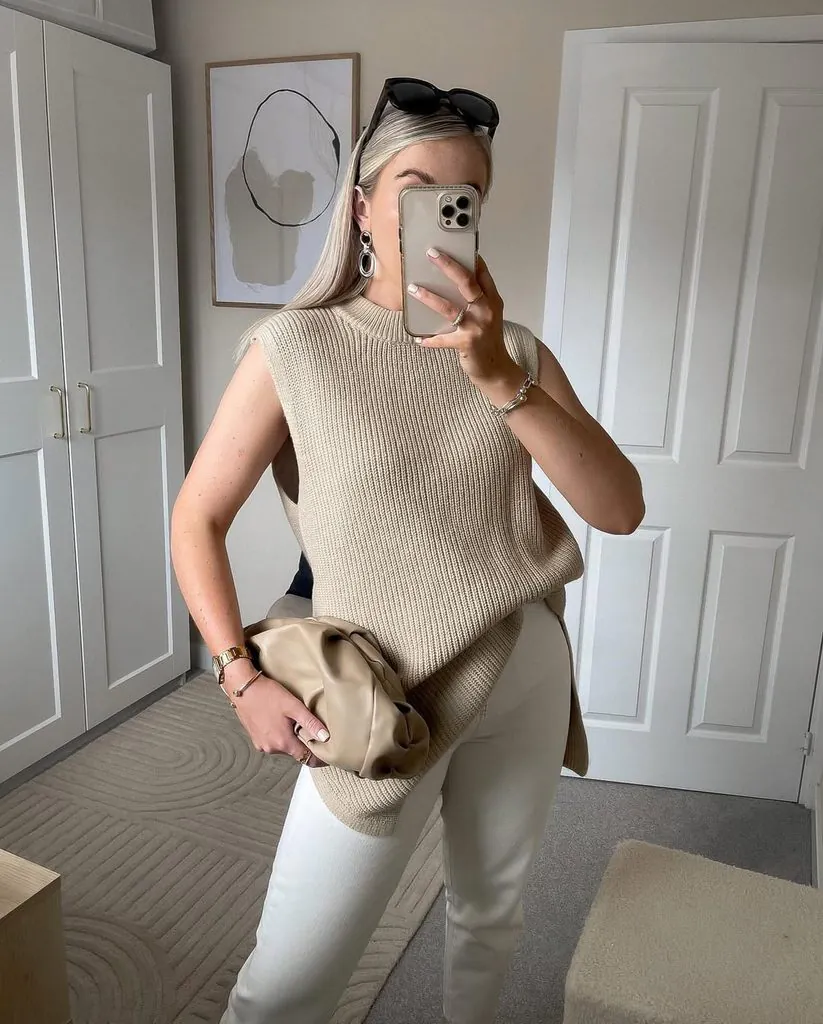 woman taking mirror selfie wearing sleeveless beige knit sweater vest and white jeans