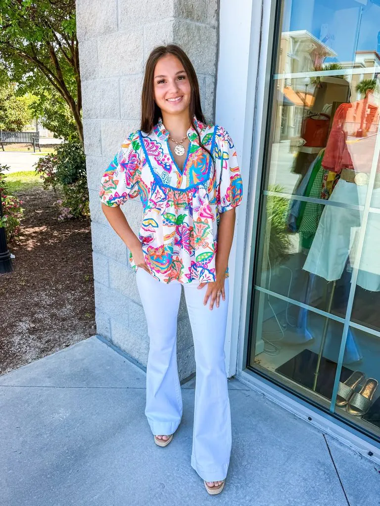 woman standing outside wearing a colorful floral pattern blouse and white jeans