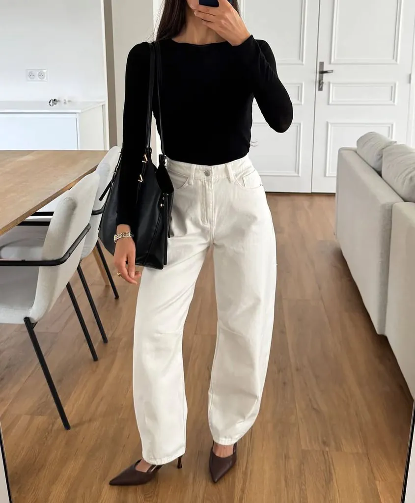 woman taking mirror selfie wearing long sleeve black top and white jeans