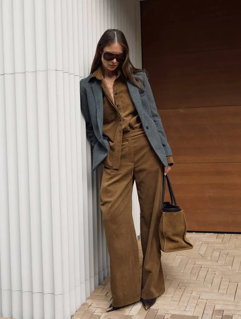 woman in grey blazer and brown corduroy suit leaning against white column