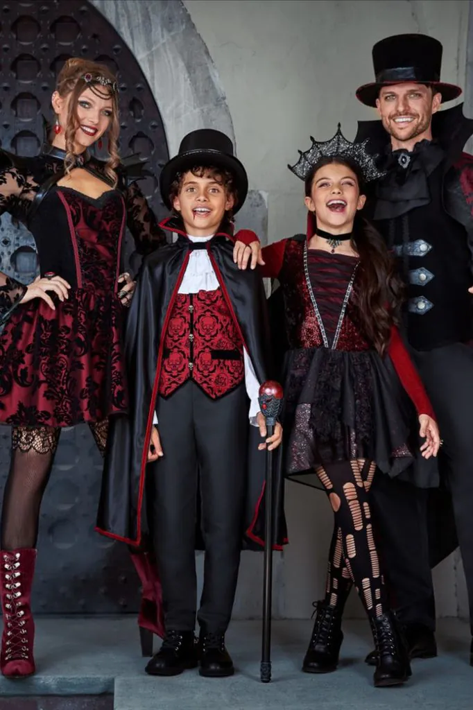 family wearing gothic vampire costumes with black cloaks posing outdoors near a door