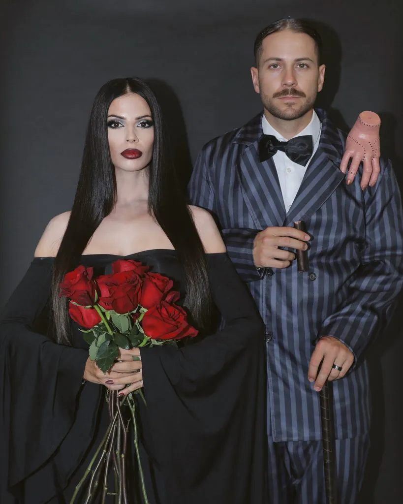 couple wearing morticia and gomez addams halloween costumes posing together in dark clothing