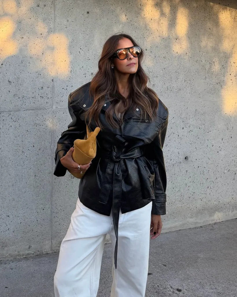 woman in belted black leather jacket white pants sunglasses holding mustard suede clutch