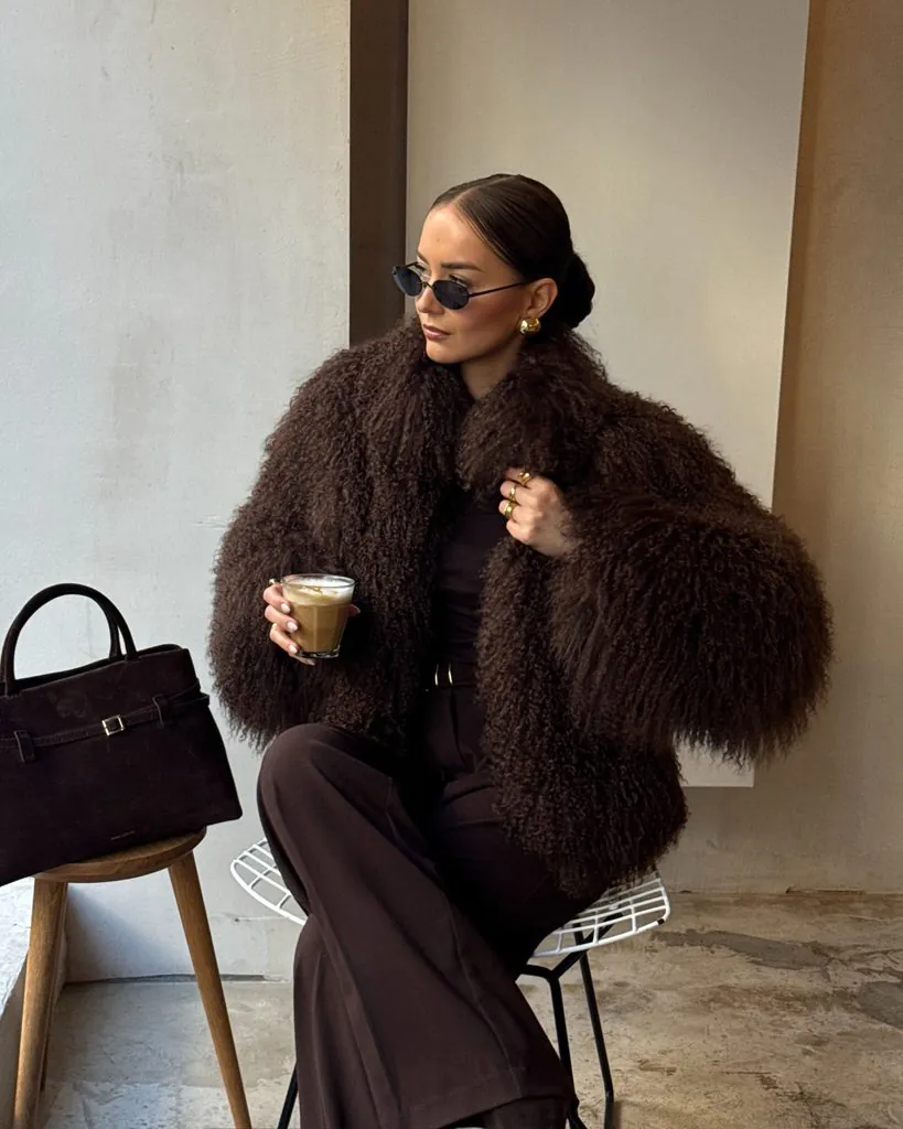 woman in dark brown faux fur jacket holding coffee cup sitting near window