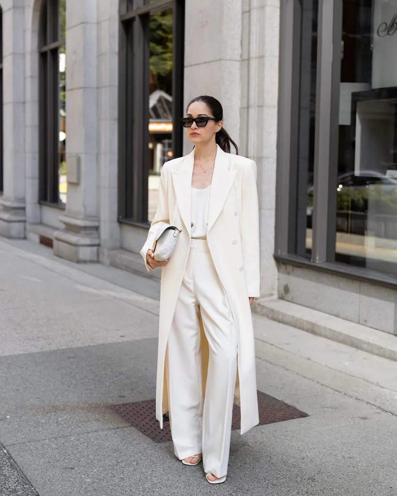 woman walking outside wearing chic all white pantsuit long trench coat and sunglasses